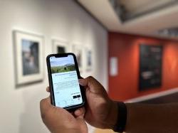 两只手托着iPhone，屏幕上显示着大学画廊(大学美术馆)的数字指南. 在后台, 在右边, 白色的墙上有相框吗, 在左边, 一面黑红相间的墙，墙上挂着假武器.